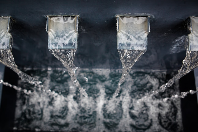 Water flows in steady streams from three metallic spouts into a basin below, creating ripples and reflections as it falls.