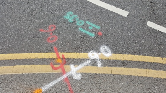 Road markings on asphalt include double yellow lines, indicating no parking restrictions, and several colored spray paint markings in red, white, cyan, and orange, possibly denoting utility lines or underground work.
