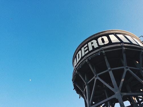 A large industrial water tower with the word 'ADERO' prominently displayed in bold white letters stands against a clear blue sky. The structure features a network of metal supports and beams.