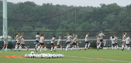 A group of people are actively warming up on a soccer field, wearing matching athletic gear. Numerous soccer balls are scattered on the grass nearby, and a lush forest background stretches across the scene behind a fence.