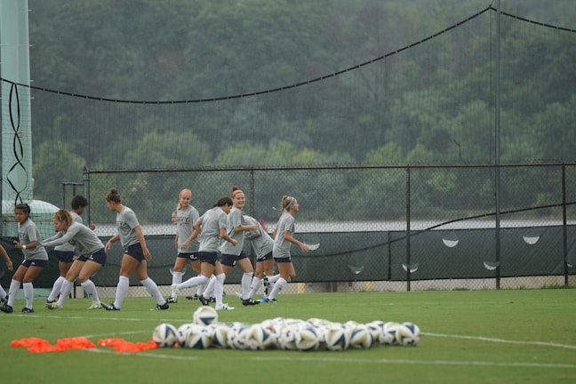 A group of female soccer players are engaged in a warm-up or training session on a grassy field. They are wearing coordinated gray shirts and shorts, with long white socks and soccer cleats. In the foreground, several soccer balls are neatly arranged on the ground. Behind them, a fence and a backdrop of lush green trees are visible, with netting also present.