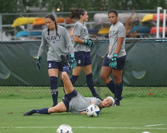 Four female soccer players are wearing grey shirts, navy shorts, and gloves. One player is on the ground, holding a soccer ball with an expression of pain or frustration, while the other three stand nearby, two looking at the player on the ground and one looking away. They are on a grassy field with a chain-link fence in the background.