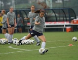 A group of women in athletic wear engages in a soccer practice on a grass field. One woman is in motion, kicking a ball, while others observe. They are wearing grey shirts with 'NAVY' printed on them, paired with navy shorts and socks. Multiple soccer balls are scattered around the field, and in the background, benches and orange cones can be seen.