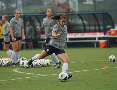 A group of women in athletic wear engages in a soccer practice on a grass field. One woman is in motion, kicking a ball, while others observe. They are wearing grey shirts with 'NAVY' printed on them, paired with navy shorts and socks. Multiple soccer balls are scattered around the field, and in the background, benches and orange cones can be seen.