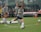A group of women in athletic wear engages in a soccer practice on a grass field. One woman is in motion, kicking a ball, while others observe. They are wearing grey shirts with 'NAVY' printed on them, paired with navy shorts and socks. Multiple soccer balls are scattered around the field, and in the background, benches and orange cones can be seen.