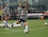 A group of women in athletic wear engages in a soccer practice on a grass field. One woman is in motion, kicking a ball, while others observe. They are wearing grey shirts with 'NAVY' printed on them, paired with navy shorts and socks. Multiple soccer balls are scattered around the field, and in the background, benches and orange cones can be seen.