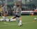 A group of women in athletic wear engages in a soccer practice on a grass field. One woman is in motion, kicking a ball, while others observe. They are wearing grey shirts with 'NAVY' printed on them, paired with navy shorts and socks. Multiple soccer balls are scattered around the field, and in the background, benches and orange cones can be seen.
