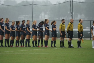 A group of soccer players in dark uniforms stand in a line on a grassy field, alongside referees in yellow shirts holding flags. The players appear ready for the start of a match under overcast skies.