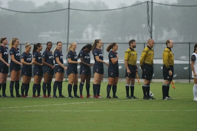 A group of soccer players in dark uniforms stand in a line on a grassy field, alongside referees in yellow shirts holding flags. The players appear ready for the start of a match under overcast skies.