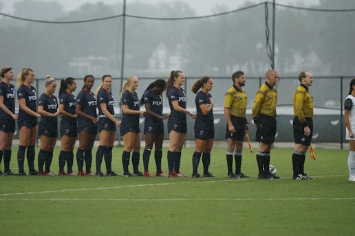 A group of soccer players in dark uniforms stand in a line on a grassy field, alongside referees in yellow shirts holding flags. The players appear ready for the start of a match under overcast skies.