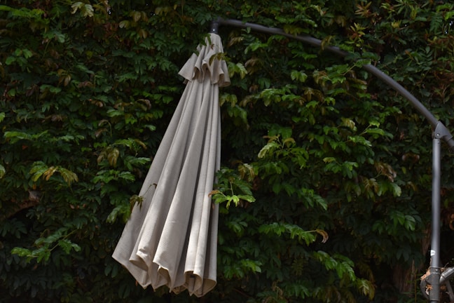 A closed beige patio umbrella hanging from a dark metal frame is positioned against a dense wall of vibrant green foliage. The leaves range from deep to lighter shades of green, adding a natural backdrop.