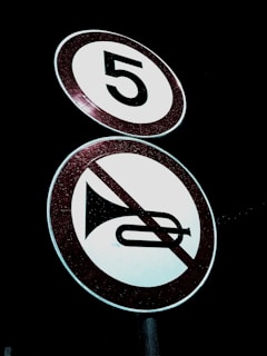 Two circular road signs are visible against a dark background. The top sign displays the number '5' indicating a speed limit. Below it, a sign features an image of a horn with a red diagonal line across it, indicating no honking.