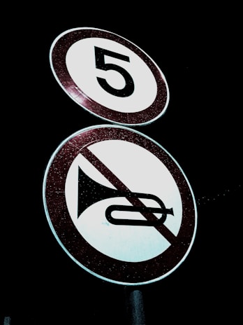 Two circular road signs are visible against a dark background. The top sign displays the number '5' indicating a speed limit. Below it, a sign features an image of a horn with a red diagonal line across it, indicating no honking.
