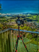 A camera mounted on a tripod is positioned on a balcony overlooking a vast landscape. The backdrop features lush green rolling hills, a patchwork of fields, and a small town with buildings scattered throughout. The sky is a vibrant blue with a few fluffy clouds on the horizon.