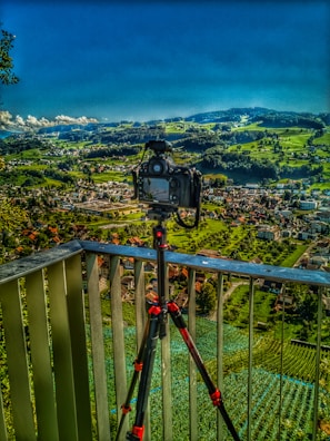 A camera mounted on a tripod is positioned on a balcony overlooking a vast landscape. The backdrop features lush green rolling hills, a patchwork of fields, and a small town with buildings scattered throughout. The sky is a vibrant blue with a few fluffy clouds on the horizon.