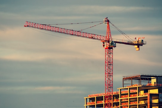 A red construction crane towers over an unfinished building, set against a cloudy sky at sunset. The crane's intricate lattice structure and extended boom stand out, with the building's concrete framework complementing the scene.