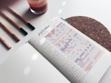 A journal or planner is open to a page labeled 'September Tracking' filled with a grid of habits tracked daily. The page includes various activities such as Pinterest, Instagram, studying, and more, with different colored markers filling the grid. To the left are three pens and a glass, while underneath is a cork coaster.