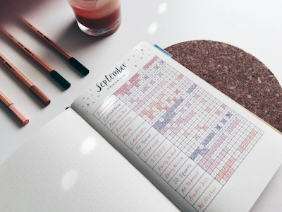 A journal or planner is open to a page labeled 'September Tracking' filled with a grid of habits tracked daily. The page includes various activities such as Pinterest, Instagram, studying, and more, with different colored markers filling the grid. To the left are three pens and a glass, while underneath is a cork coaster.