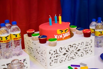 A birthday party table features a red cake with a Ninjago theme, adorned with three colorful candles. Surrounding the cake are several cupcakes with various colors of frosting. The table is decorated with intricately designed white stands, and there are multiple bottles of water with custom Ninjago-themed labels. Bright red, blue, and yellow dominate the color scheme, creating a vibrant and festive atmosphere. Plastic building blocks are also scattered on the table.