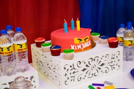 A birthday party table features a red cake with a Ninjago theme, adorned with three colorful candles. Surrounding the cake are several cupcakes with various colors of frosting. The table is decorated with intricately designed white stands, and there are multiple bottles of water with custom Ninjago-themed labels. Bright red, blue, and yellow dominate the color scheme, creating a vibrant and festive atmosphere. Plastic building blocks are also scattered on the table.