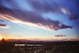 A cityscape is visible under a vibrant sky with dynamic, flowing clouds. The sunset casts a warm glow over the landscape, enhancing the silhouettes of buildings and trees. A river or body of water reflects the colors of the sky, adding a serene element to the scene.