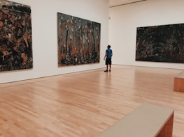 An art gallery with polished wooden floors and large abstract paintings on white walls. A person wearing a blue shirt and black shorts stands in the center, gazing at one of the artworks.