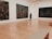 An art gallery with polished wooden floors and large abstract paintings on white walls. A person wearing a blue shirt and black shorts stands in the center, gazing at one of the artworks.