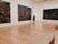 An art gallery with polished wooden floors and large abstract paintings on white walls. A person wearing a blue shirt and black shorts stands in the center, gazing at one of the artworks.