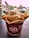 A cup of ice cream topped with a creamy scoop and sprinkled with small, pinkish pieces is served in a waffle bowl inside a branded paper cup. The cup is on a dark surface with blurred-out individuals in the background.