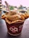 A cup of ice cream topped with a creamy scoop and sprinkled with small, pinkish pieces is served in a waffle bowl inside a branded paper cup. The cup is on a dark surface with blurred-out individuals in the background.