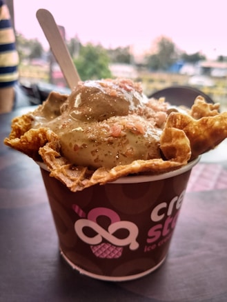 A cup of ice cream topped with a creamy scoop and sprinkled with small, pinkish pieces is served in a waffle bowl inside a branded paper cup. The cup is on a dark surface with blurred-out individuals in the background.