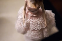 A close-up shot of a doll wearing a pink and white lace dress with intricate detailing. The focus is on the upper part of the dress, highlighting the delicate fabric and bow at the waist. The background is blurred, drawing attention to the dress and the doll's hands.