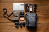 A neatly arranged collection of photography and electronic equipment on a wooden floor, including a laptop with a sticker, various cameras and lenses, memory cards, battery packs, hard drives, and a backpack. A messenger bag with a strap is also present.