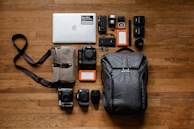A neatly arranged collection of photography and electronic equipment on a wooden floor, including a laptop with a sticker, various cameras and lenses, memory cards, battery packs, hard drives, and a backpack. A messenger bag with a strap is also present.