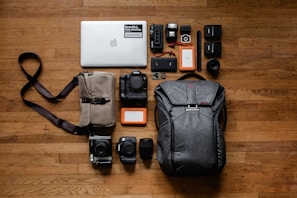 A neatly arranged collection of photography and electronic equipment on a wooden floor, including a laptop with a sticker, various cameras and lenses, memory cards, battery packs, hard drives, and a backpack. A messenger bag with a strap is also present.