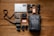 A neatly arranged collection of photography and electronic equipment on a wooden floor, including a laptop with a sticker, various cameras and lenses, memory cards, battery packs, hard drives, and a backpack. A messenger bag with a strap is also present.