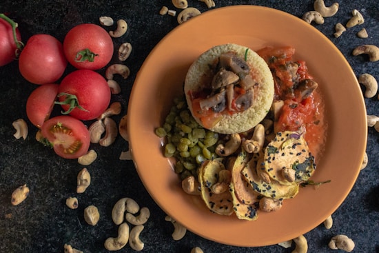 A plate with a culinary presentation consisting of couscous topped with mushrooms and tomato sauce, surrounded by peas and slices of roasted vegetables garnished with sesame seeds and cashews. The table is adorned with whole tomatoes and scattered cashew nuts.