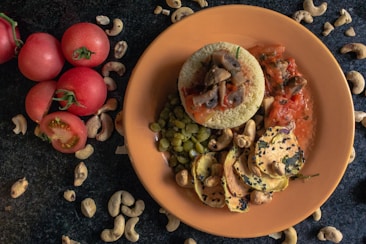A plate with a culinary presentation consisting of couscous topped with mushrooms and tomato sauce, surrounded by peas and slices of roasted vegetables garnished with sesame seeds and cashews. The table is adorned with whole tomatoes and scattered cashew nuts.