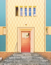 A brightly colored building facade featuring an entrance with a red door. The surrounding wall has a geometric diamond pattern in pastel yellow and white, with accents of teal and peach. Above the door are three small rectangular windows in a horizontal row. The entrance is approached via a set of gray brick stairs, and there is a metal railing on the left side.