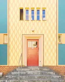 A brightly colored building facade featuring an entrance with a red door. The surrounding wall has a geometric diamond pattern in pastel yellow and white, with accents of teal and peach. Above the door are three small rectangular windows in a horizontal row. The entrance is approached via a set of gray brick stairs, and there is a metal railing on the left side.