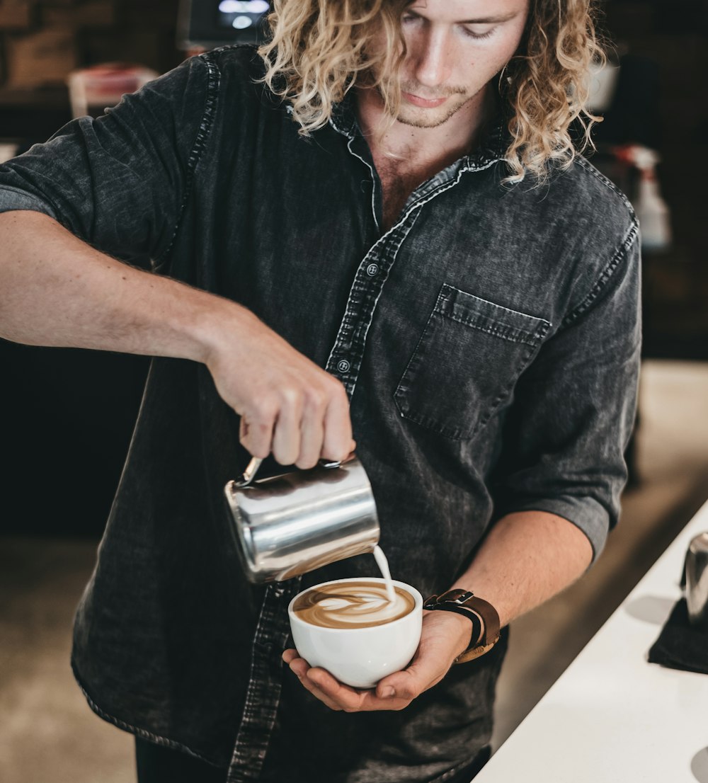 man wearing black denim jacket pouring white liquid on cup with latte art