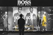 A person stands in front of a store display featuring the Hugo Boss brand. The display includes large black and white posters of models wearing suits and mannequins dressed in stylish clothing, including a yellow dress and a patterned outfit. The scene is illuminated with a focus on fashion and branding.
