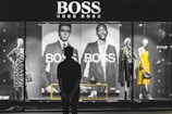 A person stands in front of a store display featuring the Hugo Boss brand. The display includes large black and white posters of models wearing suits and mannequins dressed in stylish clothing, including a yellow dress and a patterned outfit. The scene is illuminated with a focus on fashion and branding.