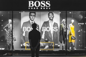 A person stands in front of a store display featuring the Hugo Boss brand. The display includes large black and white posters of models wearing suits and mannequins dressed in stylish clothing, including a yellow dress and a patterned outfit. The scene is illuminated with a focus on fashion and branding.