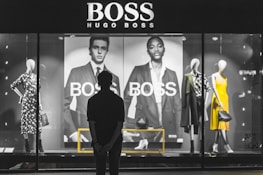 A person stands in front of a store display featuring the Hugo Boss brand. The display includes large black and white posters of models wearing suits and mannequins dressed in stylish clothing, including a yellow dress and a patterned outfit. The scene is illuminated with a focus on fashion and branding.