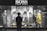 A person stands in front of a store display featuring the Hugo Boss brand. The display includes large black and white posters of models wearing suits and mannequins dressed in stylish clothing, including a yellow dress and a patterned outfit. The scene is illuminated with a focus on fashion and branding.