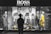 A person stands in front of a store display featuring the Hugo Boss brand. The display includes large black and white posters of models wearing suits and mannequins dressed in stylish clothing, including a yellow dress and a patterned outfit. The scene is illuminated with a focus on fashion and branding.