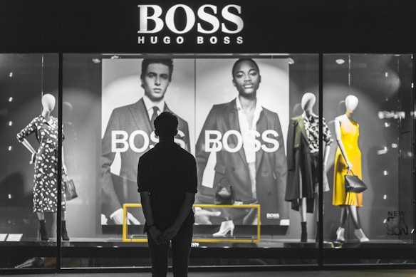 A person stands in front of a store display featuring the Hugo Boss brand. The display includes large black and white posters of models wearing suits and mannequins dressed in stylish clothing, including a yellow dress and a patterned outfit. The scene is illuminated with a focus on fashion and branding.