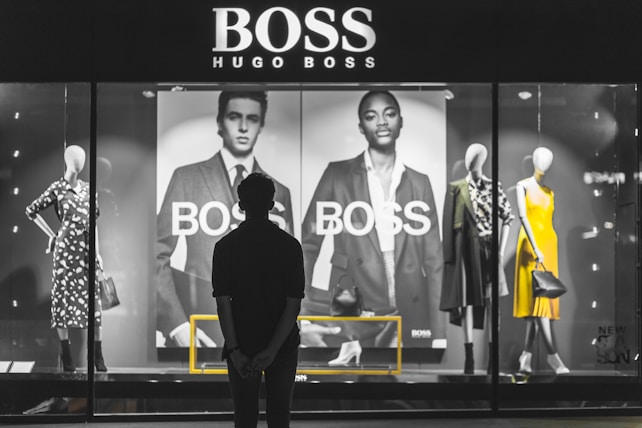 A person stands in front of a store display featuring the Hugo Boss brand. The display includes large black and white posters of models wearing suits and mannequins dressed in stylish clothing, including a yellow dress and a patterned outfit. The scene is illuminated with a focus on fashion and branding.