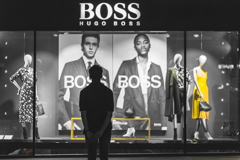 A person stands in front of a store display featuring the Hugo Boss brand. The display includes large black and white posters of models wearing suits and mannequins dressed in stylish clothing, including a yellow dress and a patterned outfit. The scene is illuminated with a focus on fashion and branding.